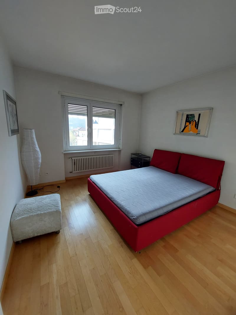 bedroom with wooden floor, white walls, red bed, white ottoman, white lamp, window with view, radiator, picture frames