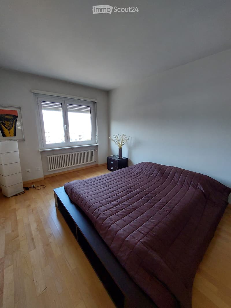 Bedroom with a large bed, wooden floor, white walls, window with blinds, nightstand, radiator, picture frame, and a cabinet.