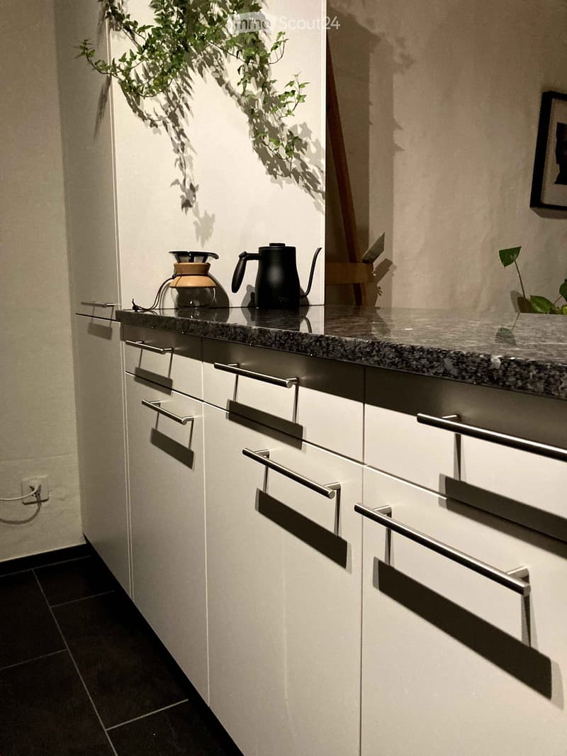 Modern kitchen with white cabinets, black granite countertop, coffee maker, and a black kettle. The kitchen has a minimalist design with sleek hardware on the cabinets.
