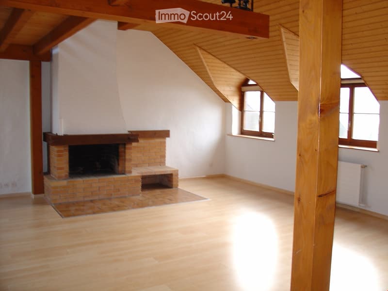 Spacious attic room with wooden beams, white walls, hardwood floors, and a brick fireplace with a mantel.