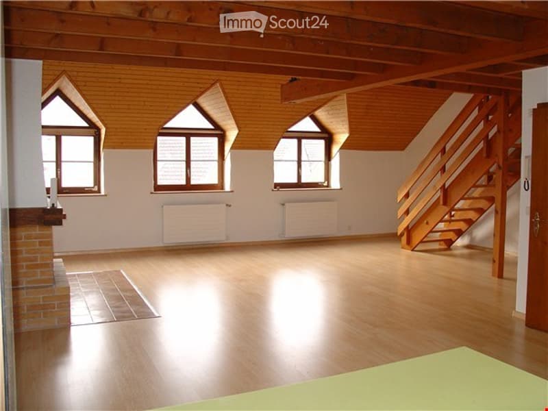 Large open space with wooden ceiling beams, hardwood flooring, and large windows providing natural light. The room appears to be a living or multipurpose area with a staircase visible.