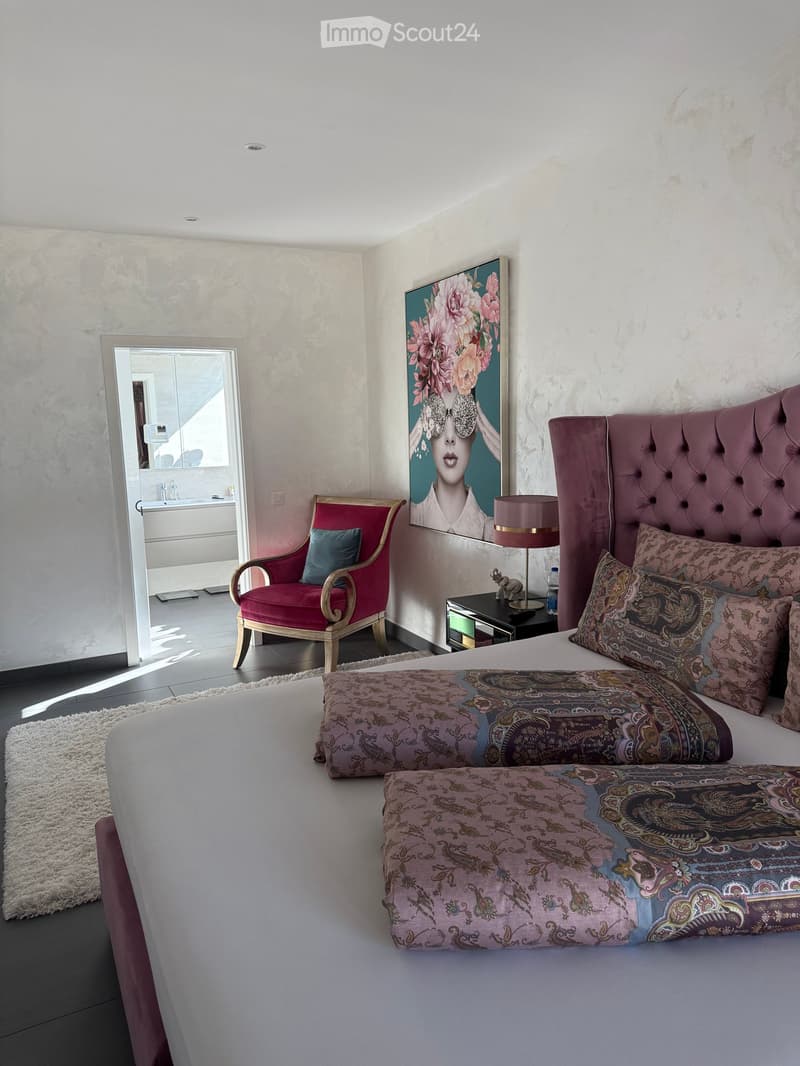 Bedroom with a large bed, a red chair, a nightstand with a lamp, a rug, and a painting on the wall. The room has a white wall and a white rug.