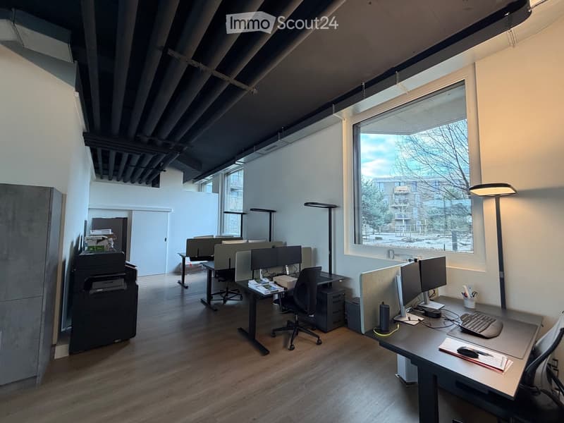 Open office plan with desks, computer monitors, office chairs, ceiling ducts, and a large window