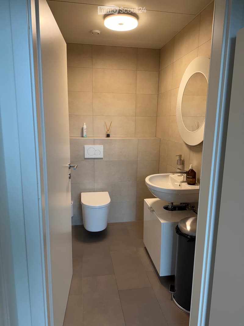 Compact bathroom with a toilet, sink, and shelves above the toilet, white tiles, circular mirror, modern sink with faucet, trash can, round light fixture
