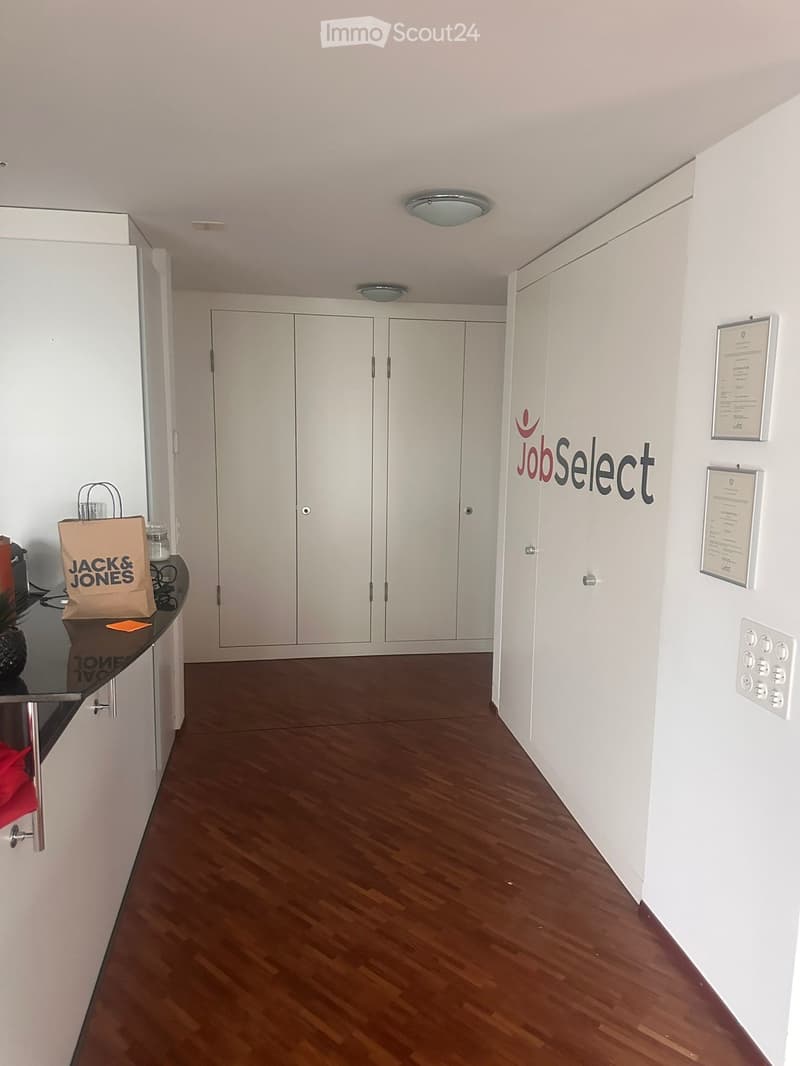 Empty room with wooden floor, white walls, white cabinets, ceiling lights, and a Jack & Jones bag on the counter.