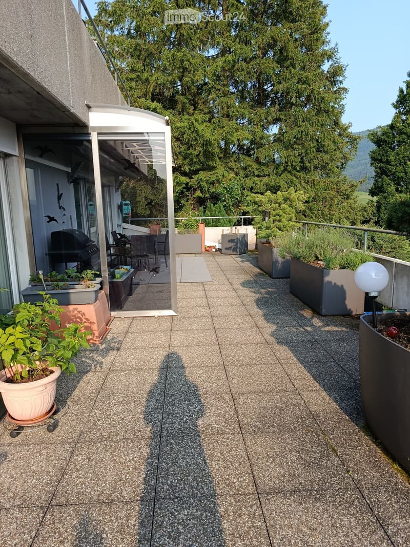 A balcony with a glass door, a table and chairs, a grill, and potted plants.