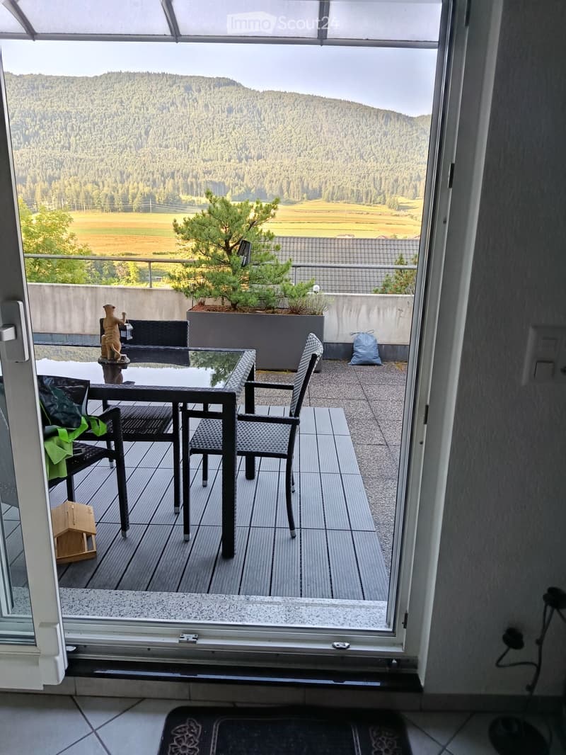 Balcony with table and chairs, potted plants, wooden floor, open door, view of mountains