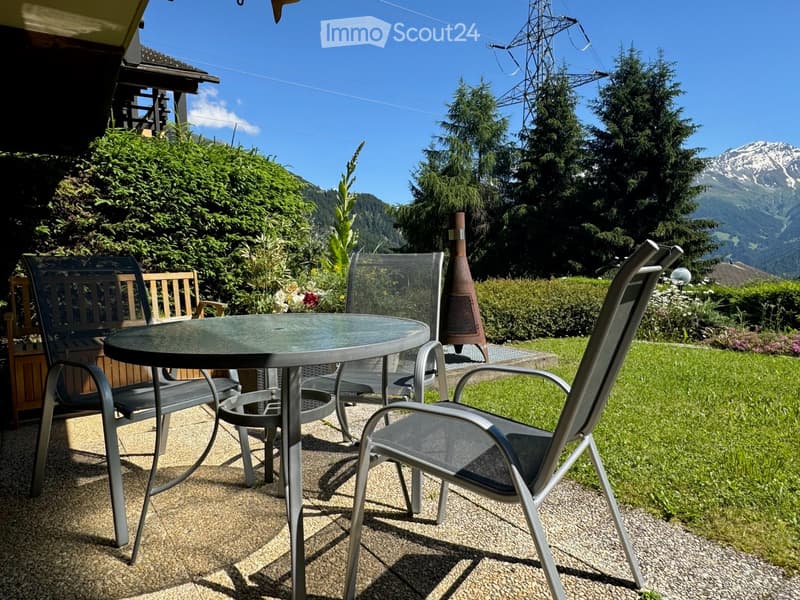 Outdoor table and chairs, wooden bench, green grass, trees, mountains in the background
