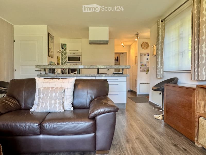 Living room with brown leather couch, white pillows, kitchen with island, white cabinets, microwave, oven, and refrigerator, wooden floor, white walls, and ceiling lights.