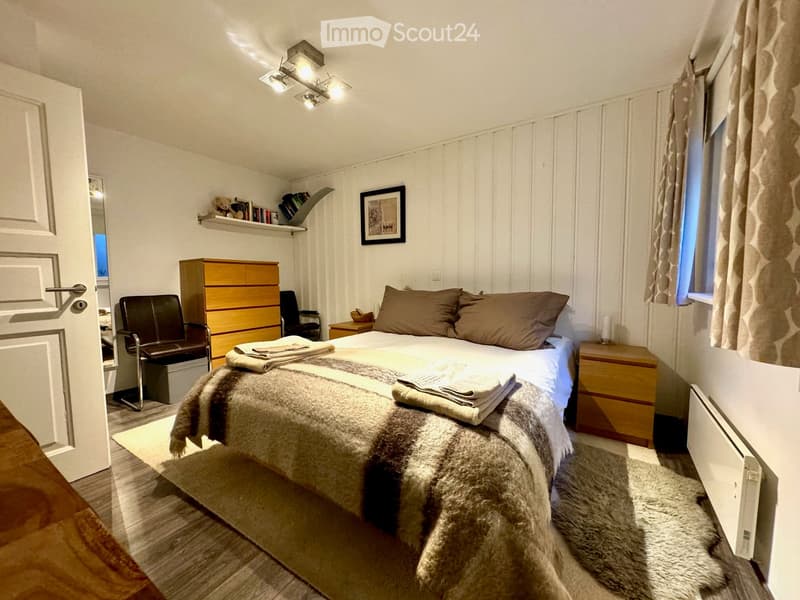 Bedroom with bed, wooden furniture, white walls, wooden floor, radiator, rug, and window with curtains