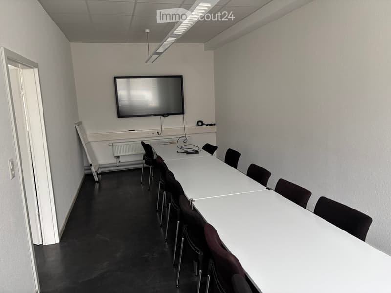 Empty room with white walls, black floor, long white tables, black chairs, mounted TV, whiteboard, radiator