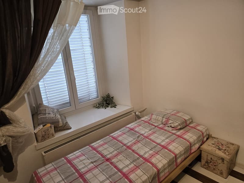 Single bed, window with white blinds, window seat with plant, storage box, pillows, single bed