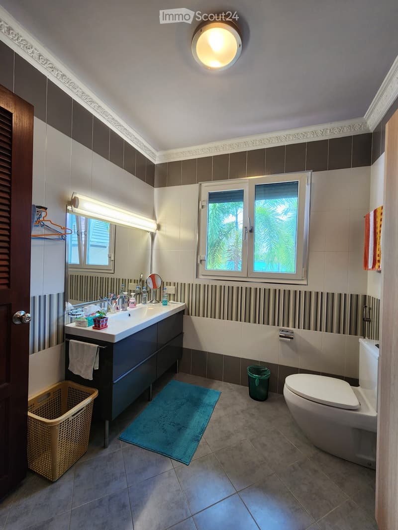 Bathroom with white sink, mirror, toilet, and window. Tiled floor and walls, with a towel hanging on the wall.