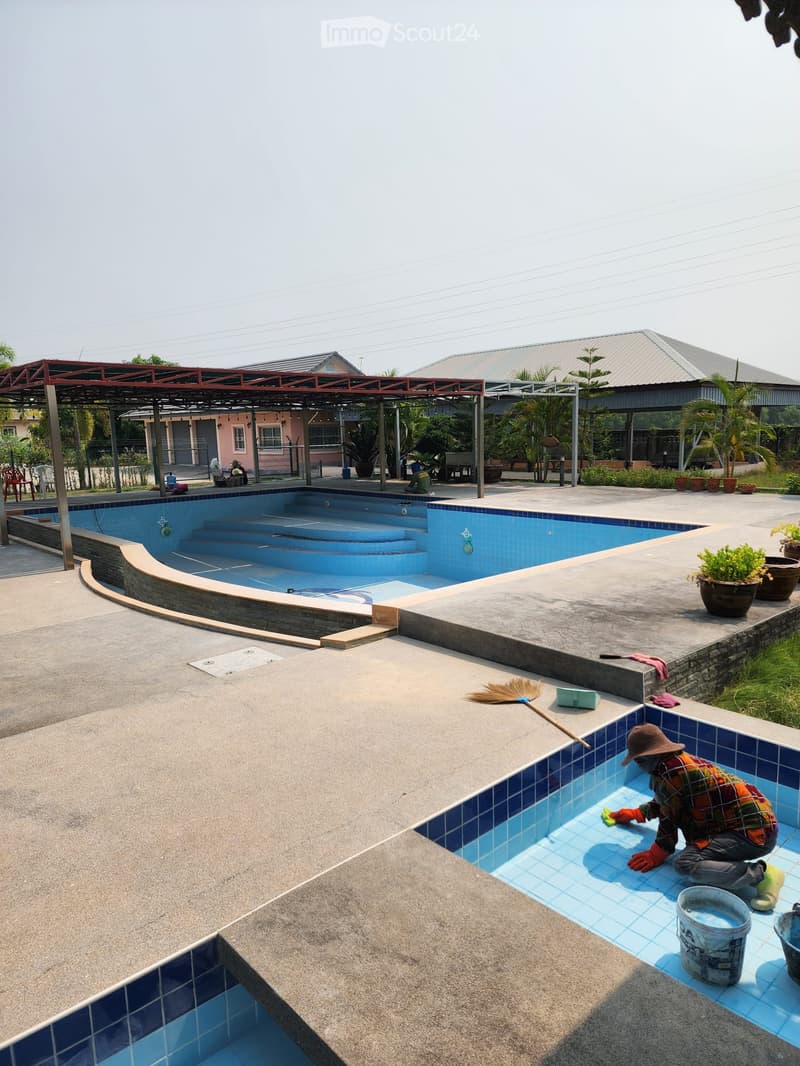 large swimming pool, tiled pool deck, covered area with metal roof, plants, person cleaning pool