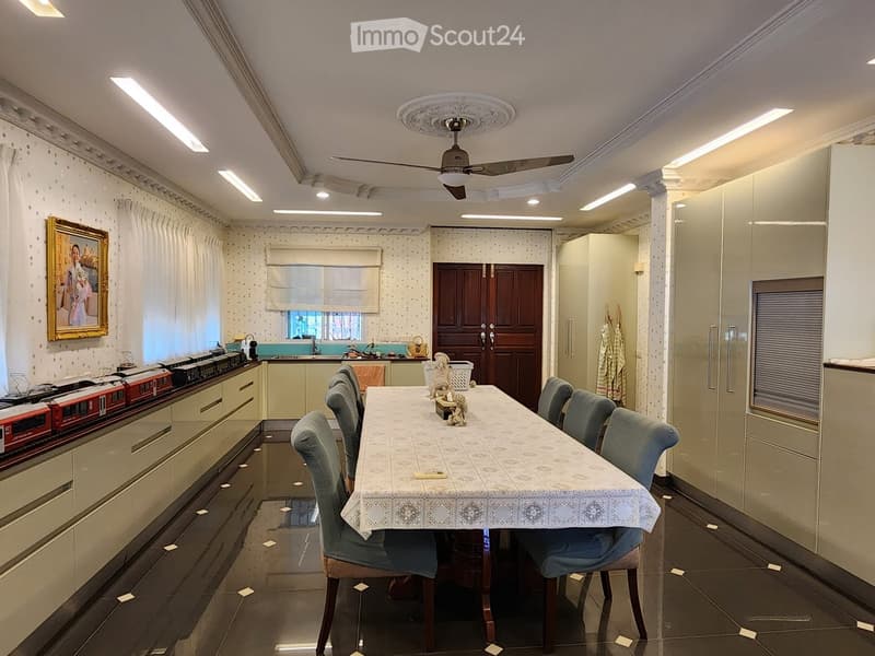 Dining room with a long table, chairs, ceiling fan, kitchen area, cabinets, picture frame, and a train set on the counter.