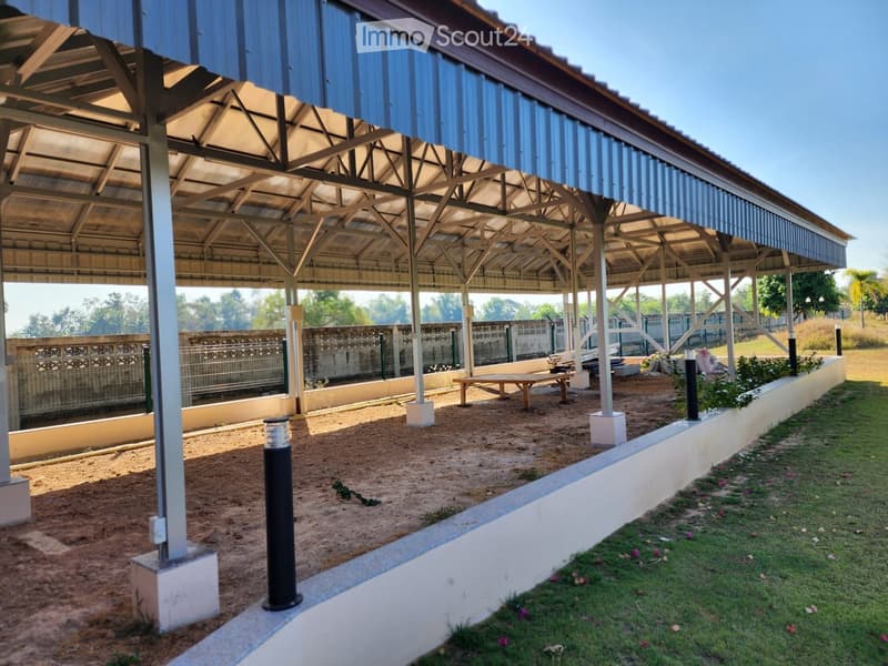 covered outdoor area with metal roof, wooden table, concrete floor, metal poles, plants, and grass