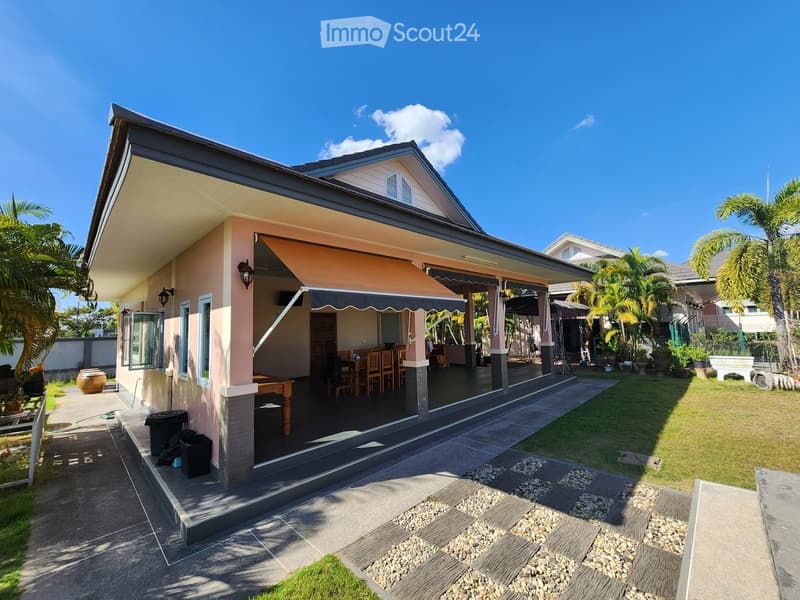 Single-story house, pink exterior, covered patio with awning, outdoor dining table, garden, trash bins, palm trees