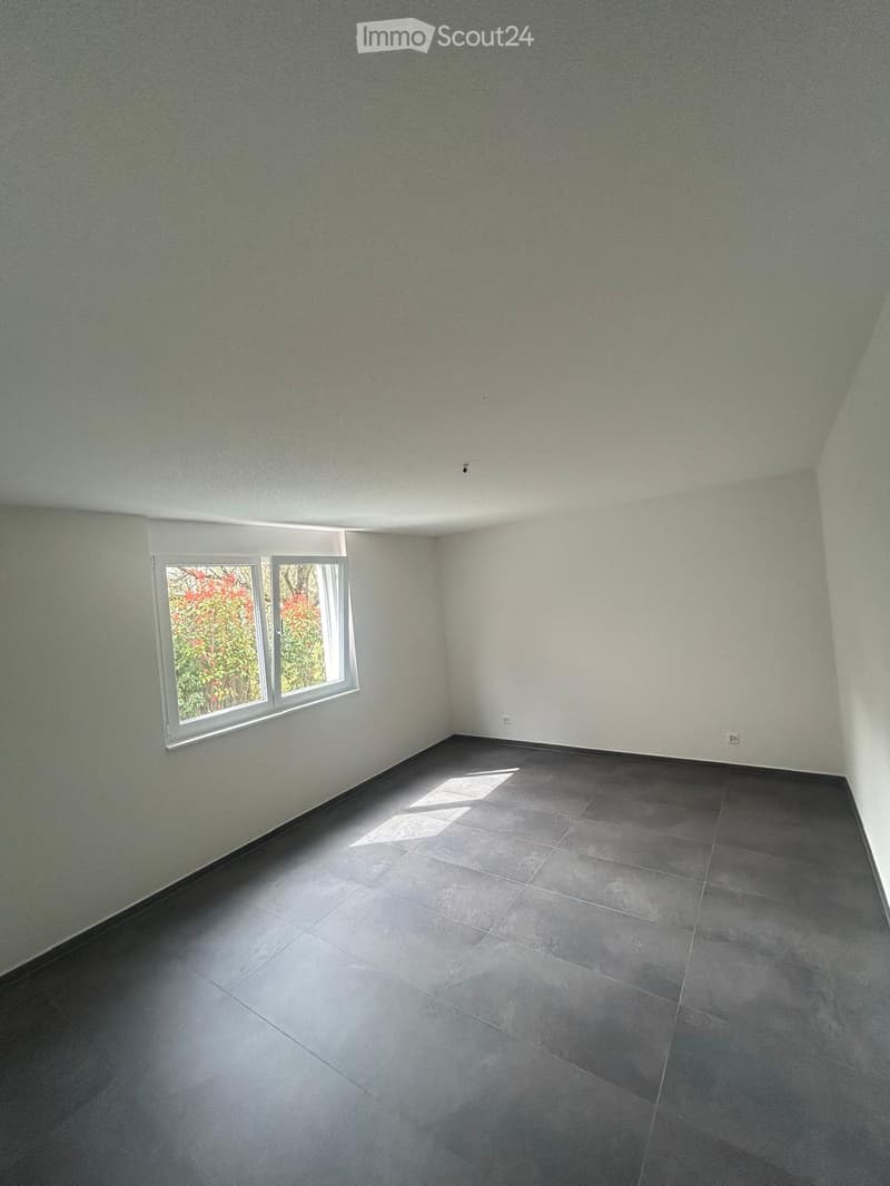 Empty room with white walls, gray floor, and a window with a view of trees outside