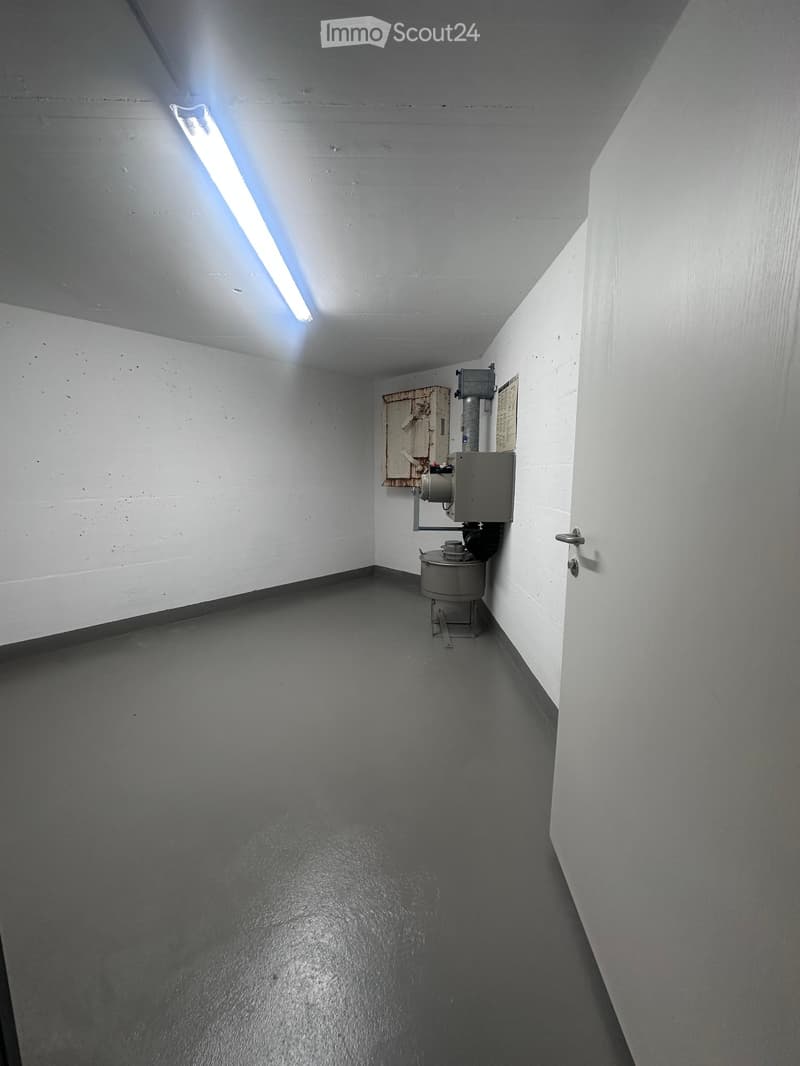 Empty room with gray flooring, white walls, a fluorescent light, a white door, and an appliance in the corner.