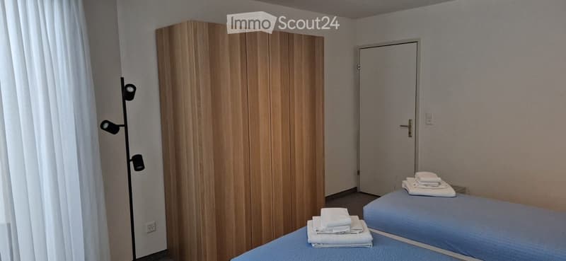 Two single beds, wooden wardrobe, white curtains, white walls, white towels, floor lamp