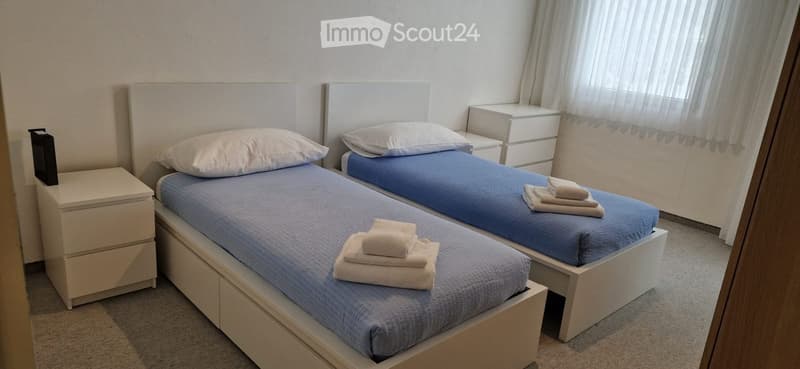 Two single beds with blue sheets, white pillows, and folded towels on top. Nightstands on both sides of the beds. A white cabinet with drawers near the window. White curtains.