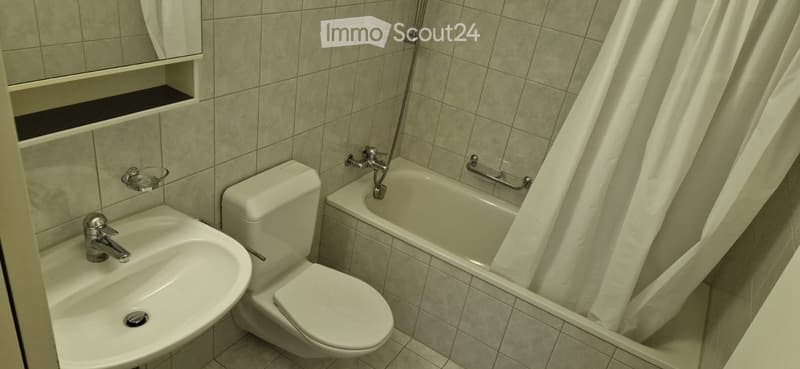 Bathroom with white tiles, white sink, white toilet, bathtub with shower curtain, mirror, and towel rack.