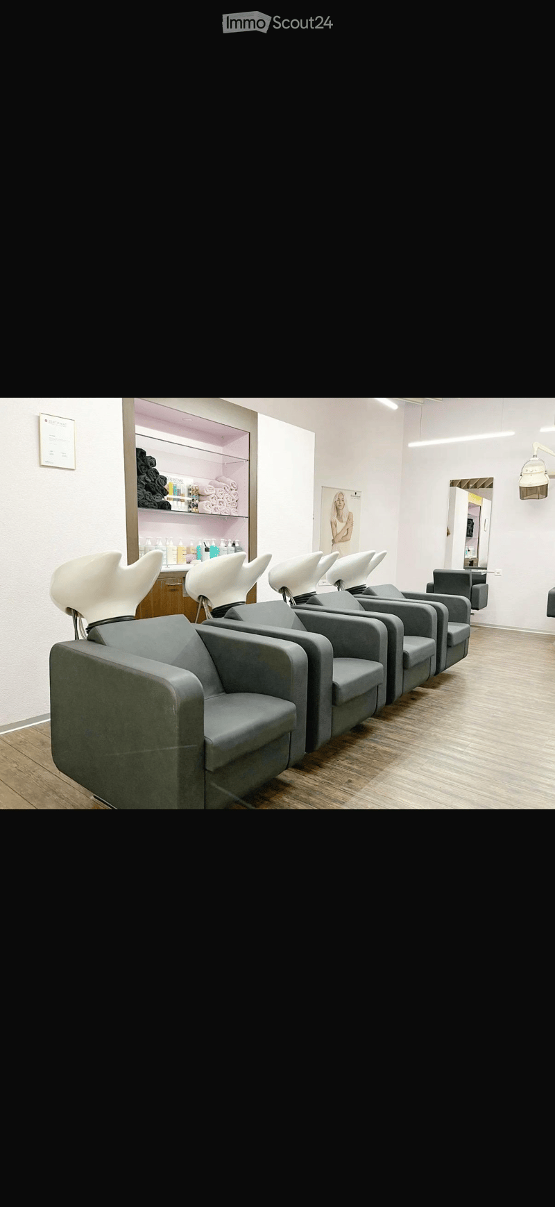 A salon with a row of grey chairs, a wooden floor, a white wall, a shelf with products, and a mirror.