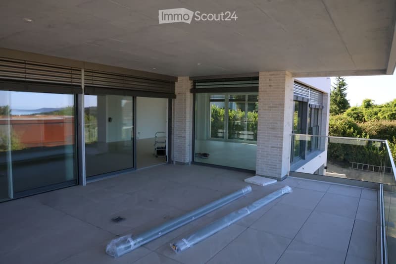 Modern balcony with large glass sliding doors, tiled floor, and a view of greenery and the sea. There are rolled-up blinds on the floor.