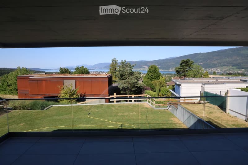 A modern balcony with a glass railing overlooks a well-maintained lawn and a scenic view of a lake and distant mountains.