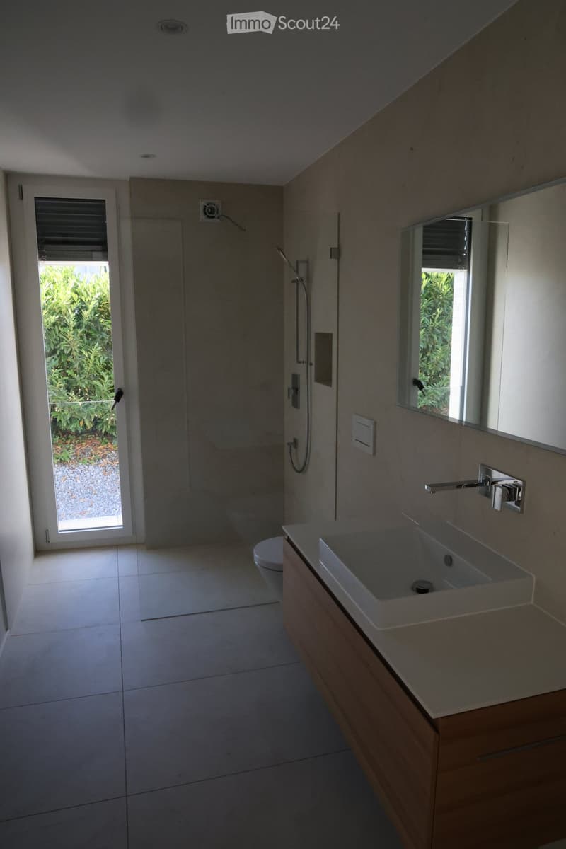 Modern bathroom with a white sink, wooden cabinet, mirror, toilet, and a glass-enclosed shower area. The room features large windows with blinds and a view of greenery outside.