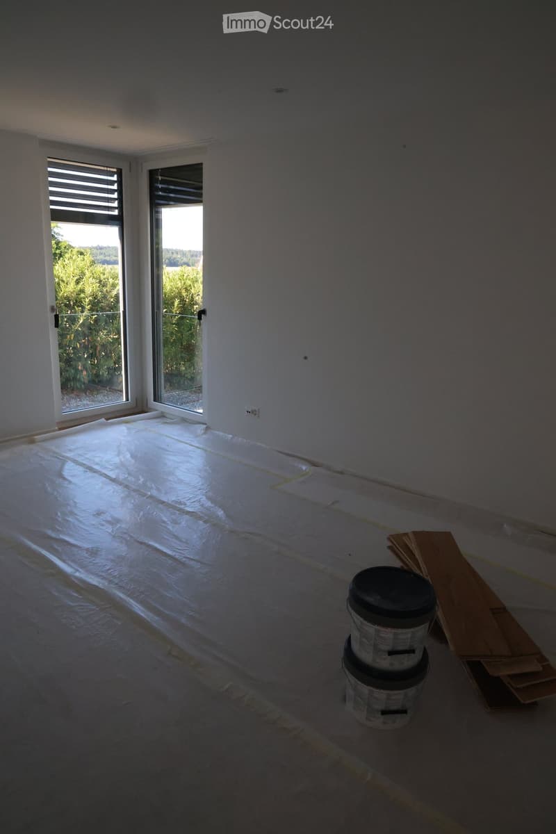 Empty room with bare walls and a plastic sheet-covered floor, featuring large windows with blinds and a view of greenery outside.