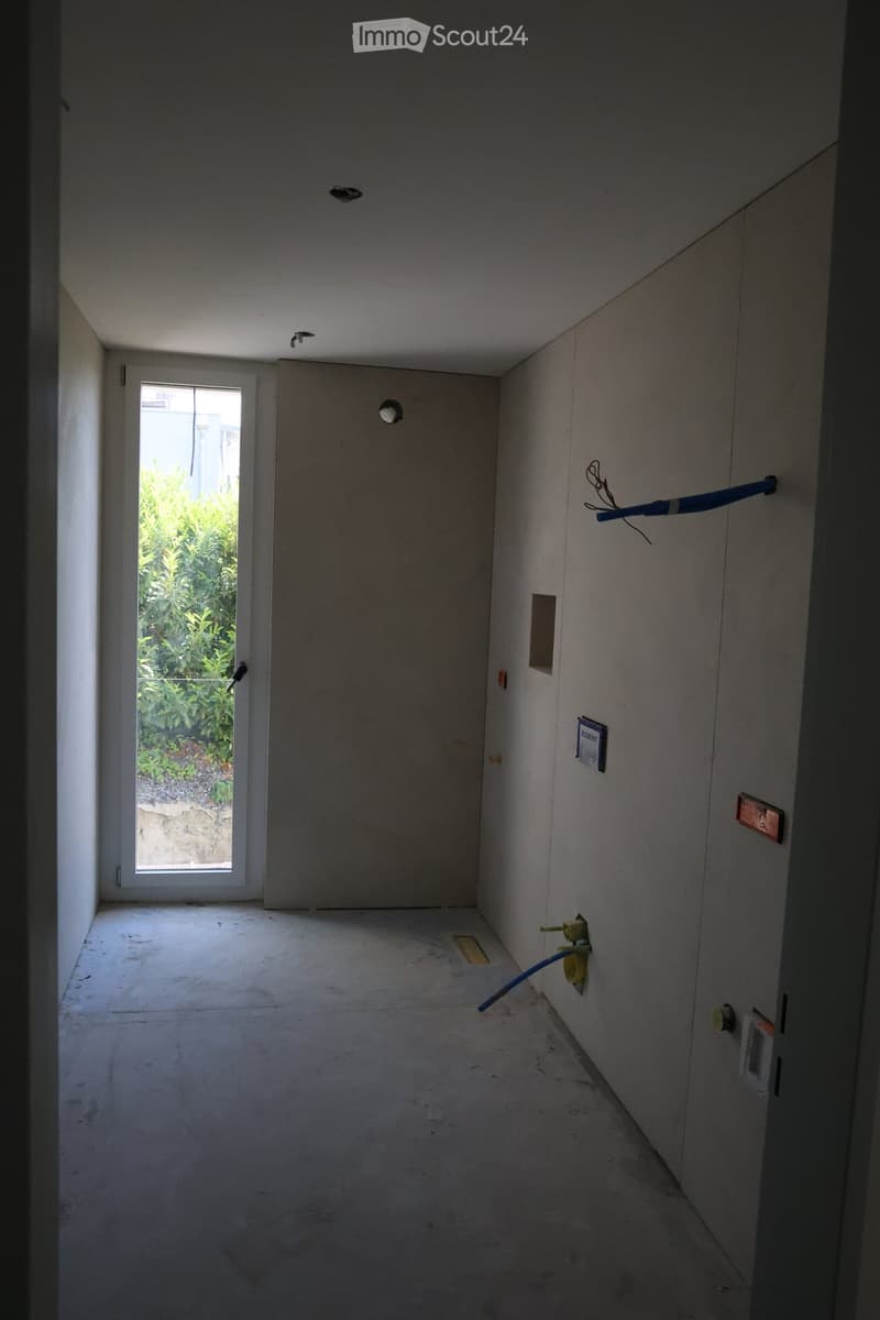 Empty hallway with concrete floor, white walls, and a window at the end showing greenery outside. Exposed electrical outlets and a blue cable are visible on the right wall.