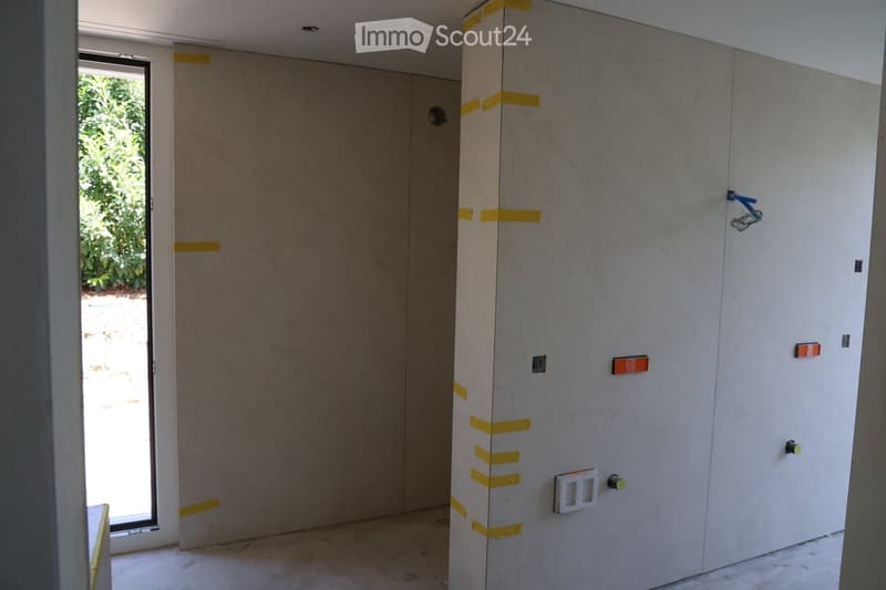 Empty room with unfinished walls, exposed concrete, and electrical outlets. Yellow tape marks the walls, and a window provides natural light.