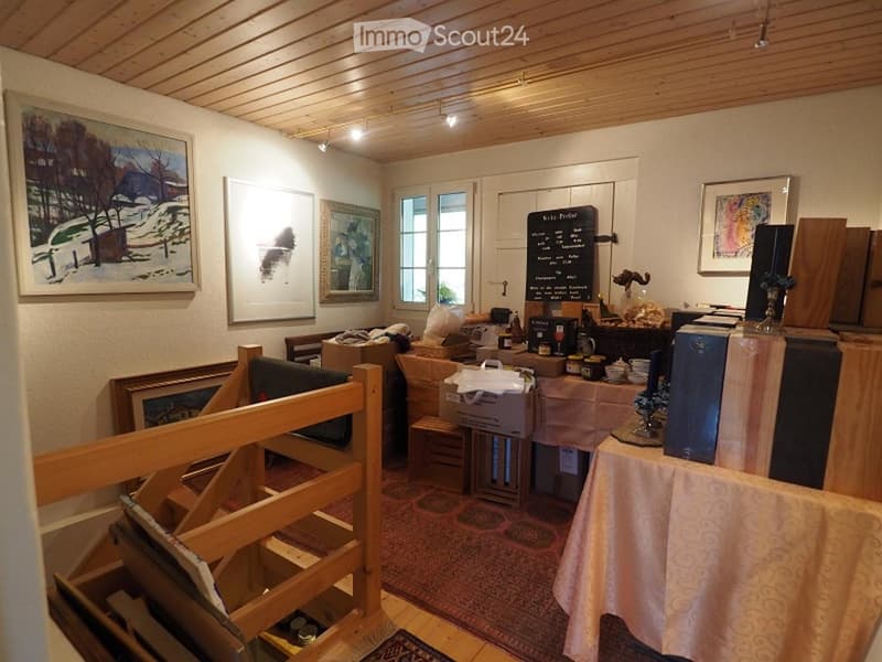 room with wooden ceiling, wooden floor, paintings on the wall, table with various items, boxes, and a rug