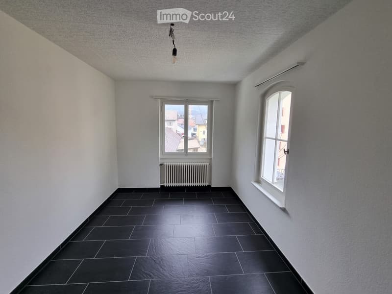 Empty room with white walls, black tiled floor, two windows, radiator, and a single light fixture on the ceiling.