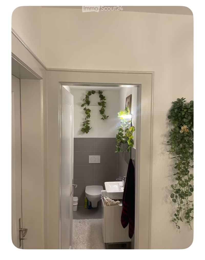 Bathroom with toilet, sink, and shower area, white walls, gray tiles, plants on the wall
