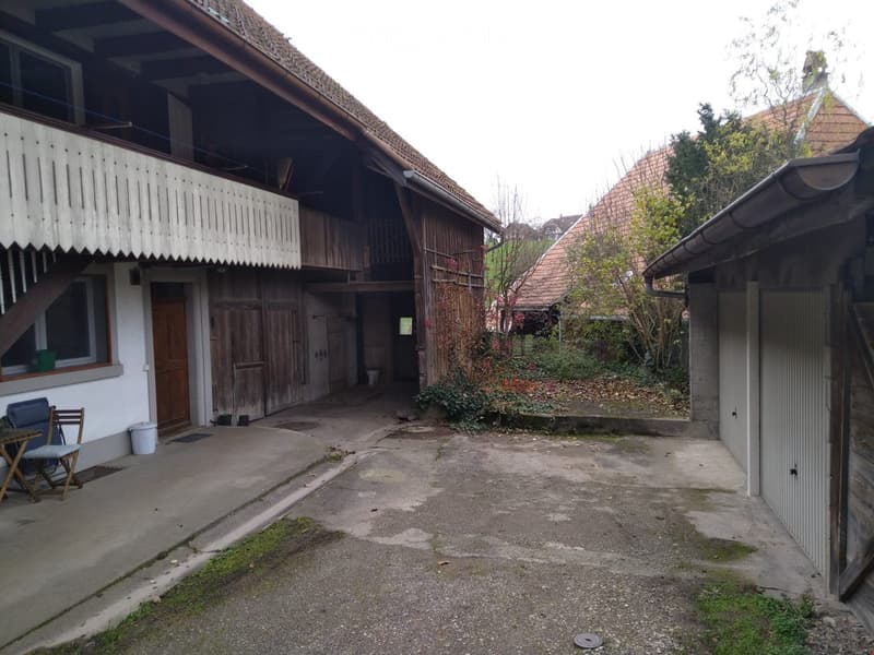 Old house with wooden doors, balcony, garage, and a small table and chair in the yard