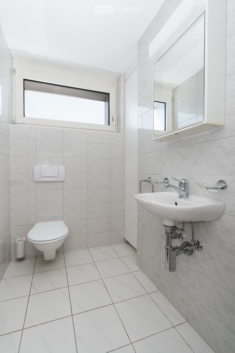 Modern bathroom with white tiles, a toilet, a sink with a chrome faucet, a mirror, and a window providing natural light.