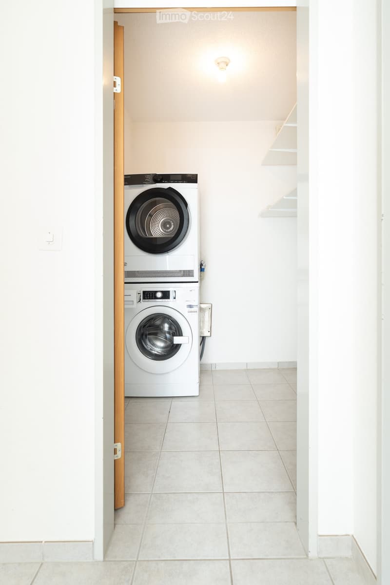 A small, clean laundry area with a white washing machine and dryer stacked vertically, white tiled floor, and white walls.