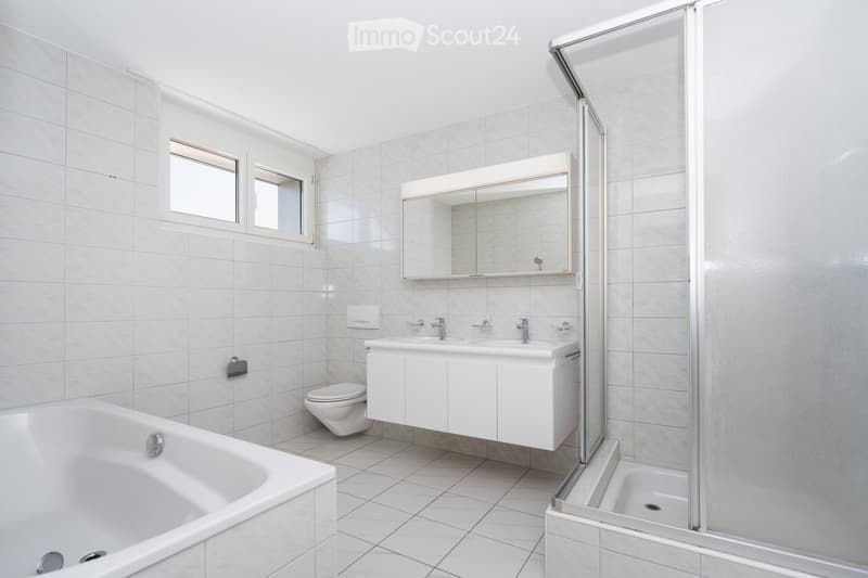 Modern bathroom with white tiles, a bathtub, toilet, double sink, large mirror, and a glass-enclosed shower area.