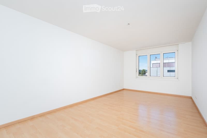 Empty room with white walls and light wooden flooring, featuring a large window with white frame and blinds.