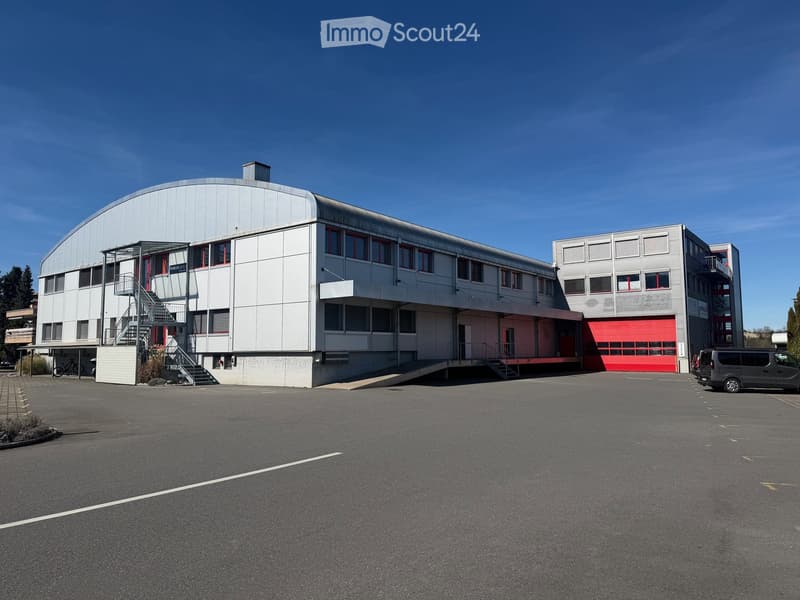 Large industrial building with white walls, red doors, and a large parking area in front