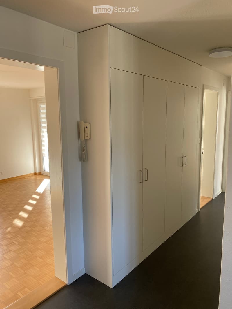 Empty room with white walls, wooden floor, large white wardrobe, and a telephone on the wall