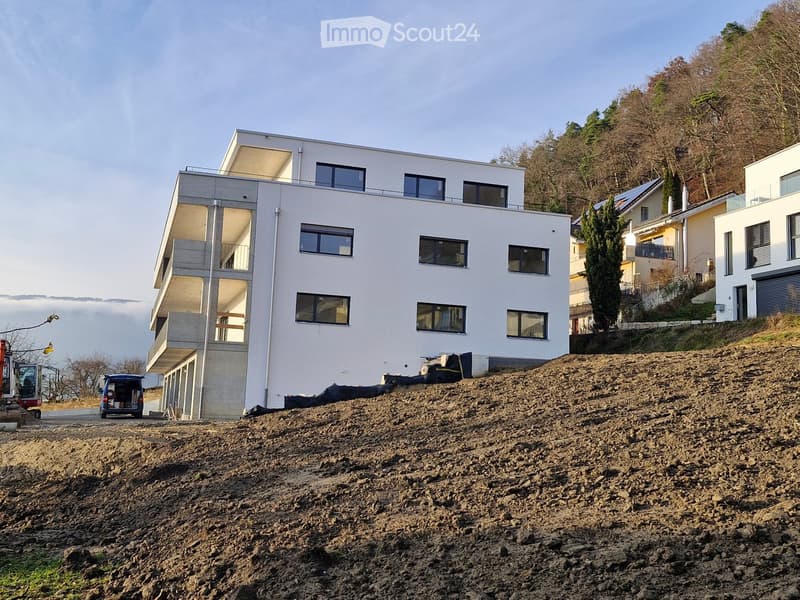Modern white building under construction, multiple windows, surrounded by trees, dirt ground