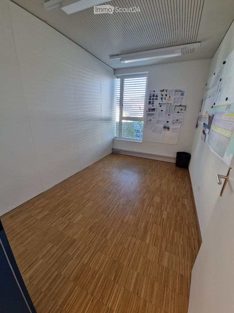 Empty room with wooden flooring, white walls, fluorescent ceiling lights, a small black trash bin, a window with closed blinds.