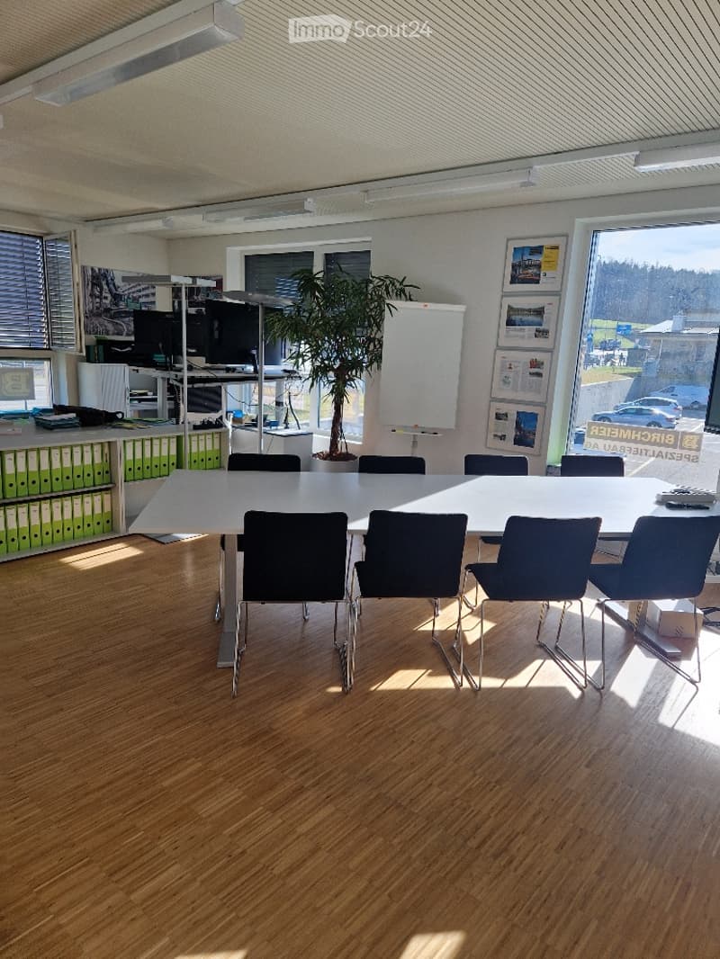 Large conference room with white walls, wooden floor, long white table with multiple chairs around, large windows, framed pictures on the wall, potted plant, and a whiteboard.