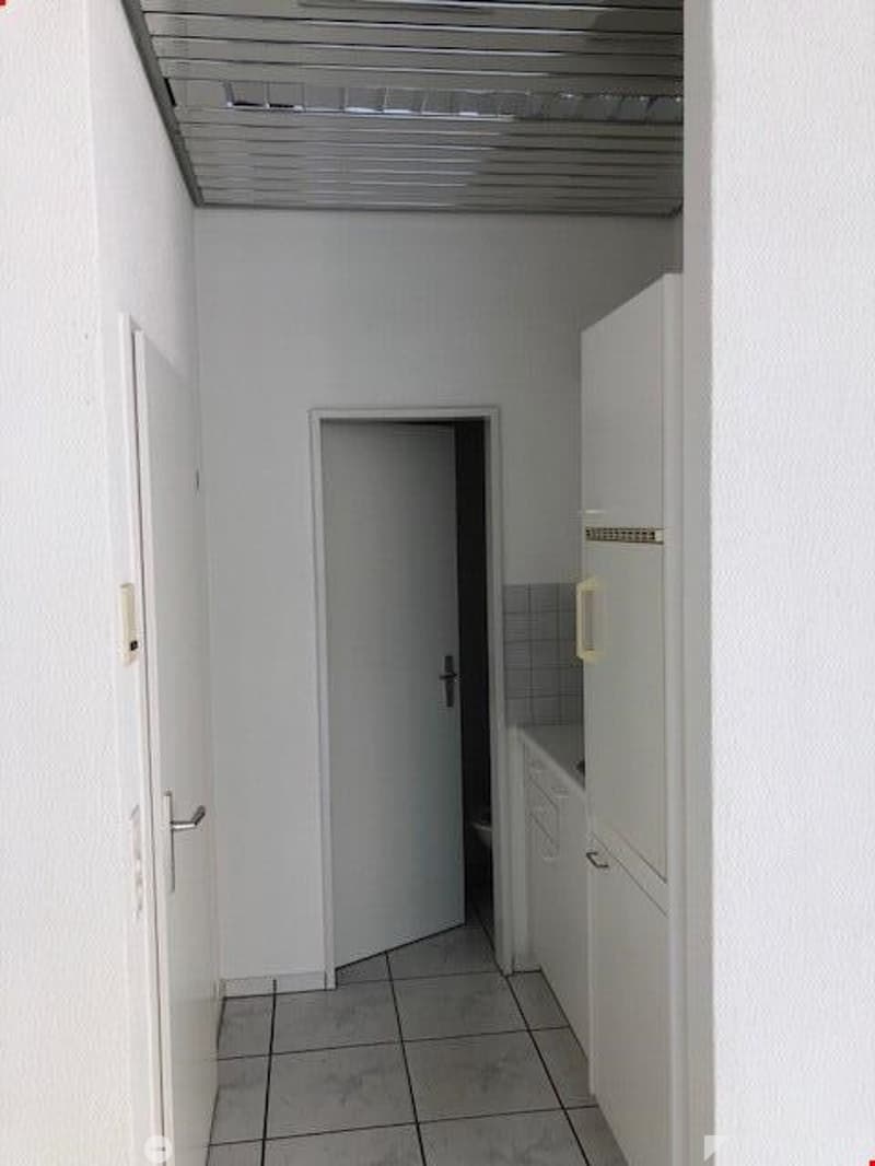Empty room with white walls, tiled floor, white door, open door leading to another room, white cabinets, and a white refrigerator.