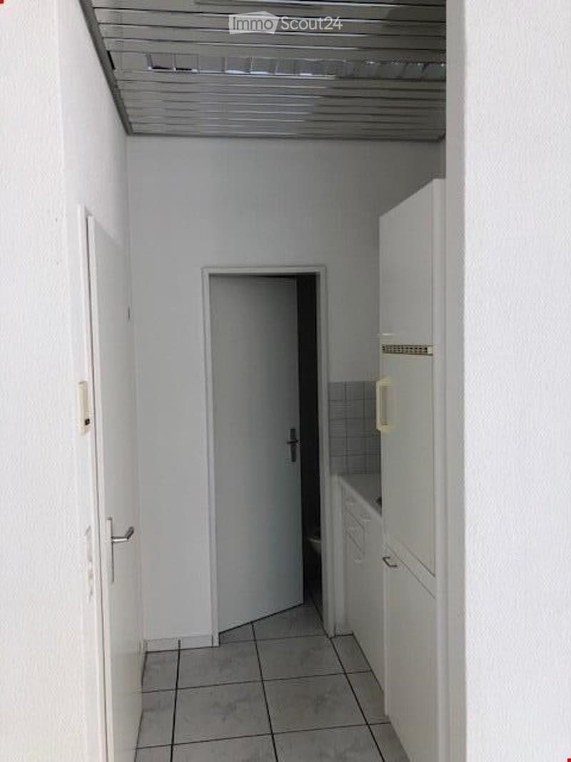 Empty room with white walls, tiled floor, white door, open door leading to another room, white cabinets, and a white refrigerator.