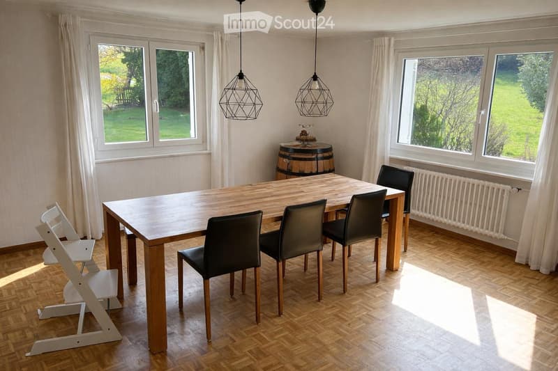 Dining room with wooden table, black chairs, white curtains, wooden floor, pendant lamps, barrel, radiator, and windows with views of a grassy field.