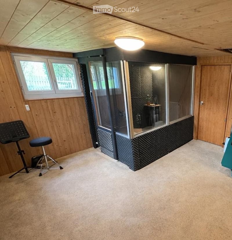 A room with wooden walls, a ceiling light, a window, a stool, a music stand, and a glass booth.